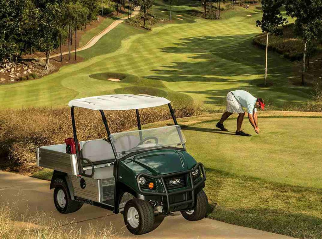 A photo of a Club Car Carryall 500 utility vehicle on a path at a golf course with a greenkeeper working on the green slightly behind it.