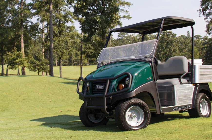 A photo of a Club Car Carryall 300 utility vehicle on a golf course with trees in the background.