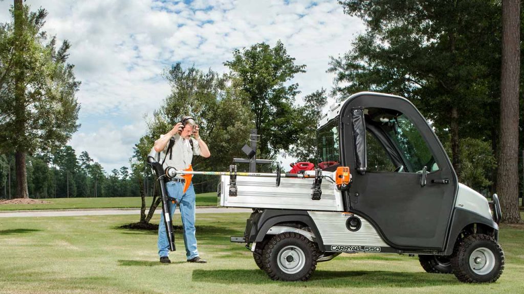 A photo of a Club Car Carryall 550 utility vehicle at a golf course with tress in the backgournd. a greenskeeper is loading equipment on the cart.